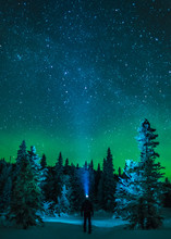 Man Standing Under Northern Lights In Winter Forest With Headlamp Beam And Starry Sky