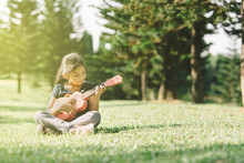 Young And Happy Asian Girl Playing With Ukelele Guitar At The Park In Sunny Morning. Hobbies And Tranquility Concept