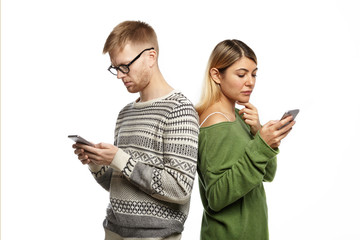 Modern technologies and internet addiction concept. Stylish young couple man and woman standing back to back, absorbed in electronic gadgets, not looking and speaking to each other, messaging online