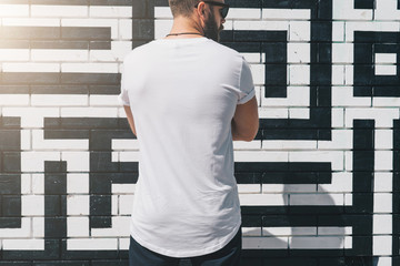 Summer day. Back view. Young bearded millennial man dressed in white t-shirt is stands against brick wall. Mock up. Space for logo, text, image. Instagram filter, film effect, bokeh effect.