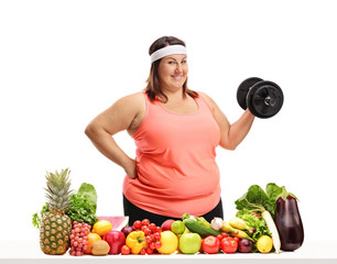 Wall Mural - Overweight woman holding a dumbbell behind a table full of fruit and vegetables