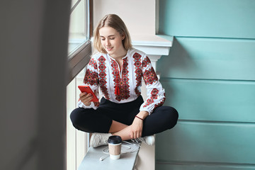 Young beautiful caucasian girl student wearing a vyshyvanka, a traditional Ukrainian embroidered shirt checks her smartphone while drinking coffee between lectures