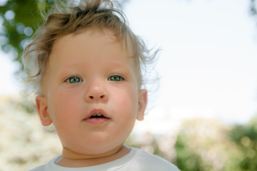 a little curly boy in a white T-shirt looks directly at me, in the street in the summer, close-up