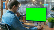 © Gorodenkoff - Young Man Works at His Desk on the Personal Computer with Mock-up Green Screen. In the Background His Colleague Works in the Creative Office.