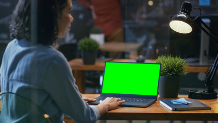 Poster - Female Office Worker at Her Desk Works on a Laptop with Mock-up Green Screen. Over the Shoulder Footage. She sits at the Wooden Desk in Creative Office.