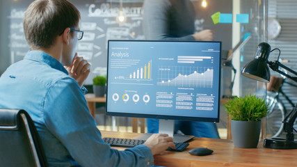 Poster - Analyst Works on a Personal Computer Showing Statistics, Graphs and Charts. In the Background His Coworker and Creative Office.