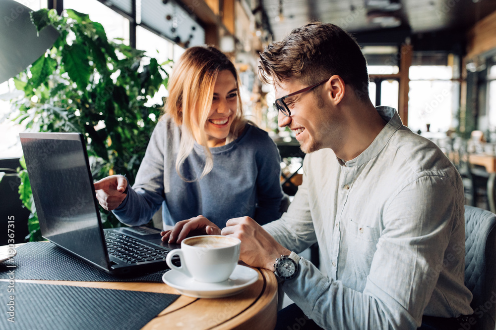 Attractive man in eyeglasses and charming woman is pointing at the laptop screen, laughing together, resting at cafe with cup of coffee. - obrazy, fototapety, plakaty 