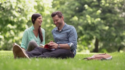 handheld shot of playful girlfriend sitting with boyfriend holding book on field at park