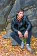 © Alexander Image - American man autumn/winter casual street fashion. Young handsome man wearing black leather jacket, pants, gray leather shoes, sits on rocks with fallen leaves on ground in Central Park, New York..