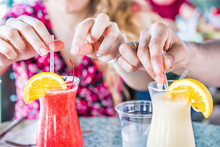Closeup Of Two Alcoholic Summer Drinks, Strawberry Daiquiri And Pina Colada, In Restaurant With People's Hands Touching, Taking Straws