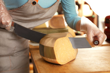 Wall Mural - Young woman cutting delicious cheese with dutch knife on table in shop