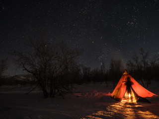 a night photo of arctic landscape with a tent and a silhouette of a person illuminated by a fire in 