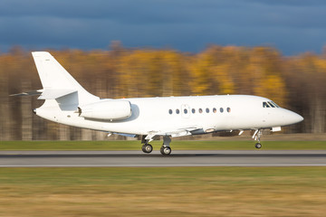 Side view of a luxury corporate or private jet landing in motion/ Landing business jet low over the runway, day light, blurred autumn forest on background/ 