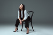 full length view of beautiful young asian woman sitting on chair and looking at camera on grey