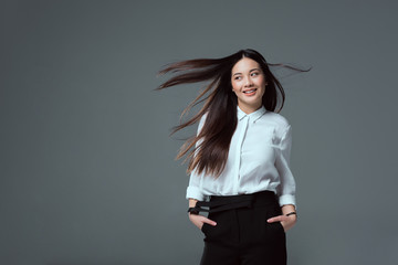 Wall Mural - beautiful smiling asian girl standing with hands in pockets and looking away isolated on grey