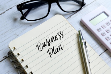 Sunglasses,calculator,pen and notebook written 'BUSINESS PLAN' on white wooden background.