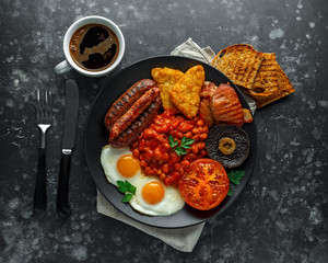 Full English breakfast with bacon, sausage, fried egg, baked beans, hash browns and mushrooms in black plate. cup coffee.