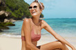 © WHstudio Leushin N - Young happy female wears sunglasses, headband and swimsuit, poses against sea cliff and beautiful blue water, has vacations on exotic beach, poses for travel magazine. Summer and leisure concept