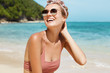 © WHstudio Leushin N - Positive pleased female model spends summer holidays on exotic tropical sea shore with cliff behind, laughs joyfully as recieves pleasant compliment about her perfect slim body. Lifestyle and rest