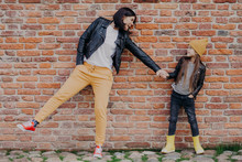 Small Fashionable Girl And Her Mother Have Fun Together, Keep Hands Together, Pose Against Brick Wall. Stylish Young Mum And Daughter Play Outdoor. Relationships, Good Mood And Fashion Concept
