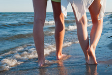 Romantic Date Male And Female Legs Walking Along The Seashore, Waves Splashing Water Foam Beach At Sunset
