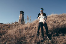 Young Loving Couple Of Teenagers Standing On The Hillside Against The Background Of The Old Lighthouse And Looking Into The Distance.