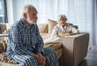 Upset old man sitting on bed with sad look. His woman sitting in bed under the blanket and looking at him with sympathy