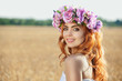 Beautiful red-haired woman in a flower wreath in a wheat field in summer at sunset.