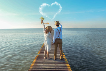 couple from behind with vintage dress holding hands stand on bridge looking at heart shaped cloud on bright blue sky,romantic atmosphere