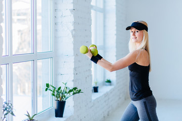 Wall Mural - Side view of blond slim fitness woman workout with dumbbells in front of window in gim studio loft interior. Young girl holding sport equipment.