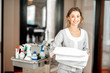 Portrait of a young woman chambermaid holding a towel standing with maid cart full of cleaning stuff in the hotel corridor