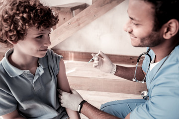 Wall Mural - Brave patient. Side view on relaxed teenager and a physician having pleasant chat and smiling before vaccination indoors.