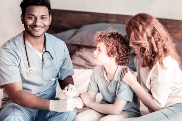 Wall Mural - Always ready to help. Cheerful male medical worker grinning broadly into the camera while putting a bandage on an arm of a young patient.