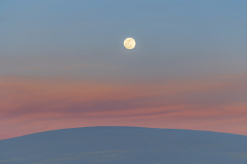 a super moon rises over sand dunes and the remnants of sunset lit clouds