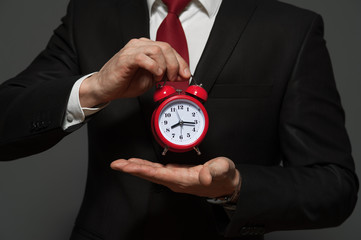 Wall Mural - Just in time concept. Close-up of young businessman holding red clock in hands on gray background