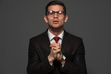 Wall Mural - Portrait of middle-aged businessman in formal suit and glasses with crossed hand near his chest and praying against gray background