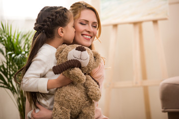 Wall Mural - cute little child holding teddy bear and kissing happy mother at home