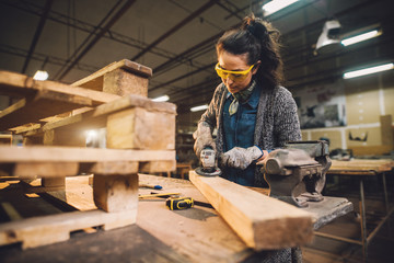 Portrait view of hardworking middle aged professional female carpenter worker working with sandpaper and choosing wood in the workshop.