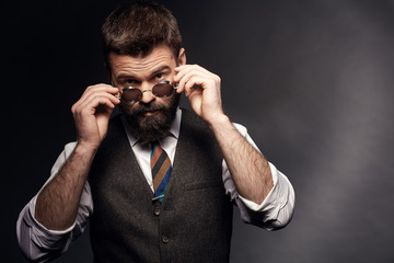 Studio portrait of attractive handsome man with dark hair, mustache and beard in white shirt, brown vest and colorful tie takes off round sunglasses