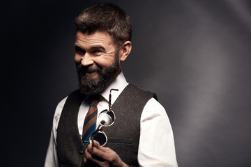 Wall Mural - Studio portrait of smiling attractive man with dark hair and mustache in white shirt, brown vest, colorful tie and glasses in hand. Old fashioned style, gentleman.