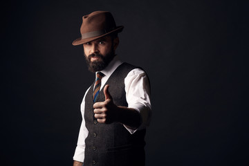 Wall Mural - Studio portrait of handsome man with dark hair and mustache in white shirt, brown vest, colorful tie, brown hat with thumb up. Old fashioned style, gentleman.