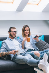 Wall Mural - Young couple sitting on the couch, drinking coffee and watching tv  