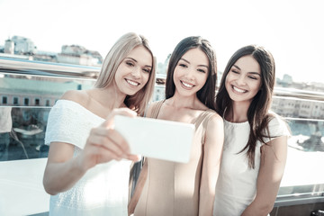 Wall Mural - Beautiful photo. Happy positive joyful woman smiling and taking a selfie while standing together with her friends on a rooftop