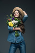 © LIGHTFIELD STUDIOS - attractive smiling girl with closed eyes in denim clothes and straw hat holding bouquet of flowers, isolated on grey