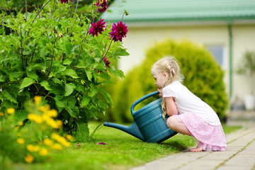 Wall Mural - Cute little girl watering flowers in the garden at summer day. Child using garden hose on sunny day