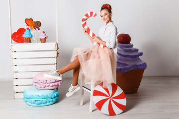 Beautiful teenage model girl in pink tutu or tulle skirts in studio on white backgroud with big artifitial macaroons, fancy cakes, candies and sweets