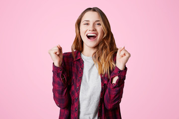 Waist up portrait of overjoyed woman in checkered shirt, clenches fists, rejoices great success and achievement aims, isolated over pink background. Pleasant looking young happy female gestures