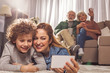 Portrait of happy female and smiling kid taking selfie by mobile while locating on carpet. Beaming grandparents drinking cup of tea and sitting on sofa. Entertainment concept