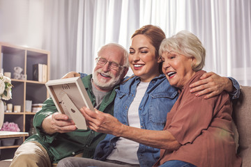 Wall Mural - Portrait of laughing grandparents and smiling young woman looking at photo in room. Remembrance concept