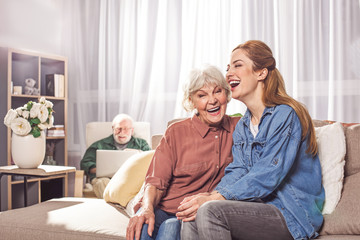 Wall Mural - Portrait of cheerful old woman laughing with young lady on sofa. Beaming bearded grandfather typing in laptop. Happiness concept